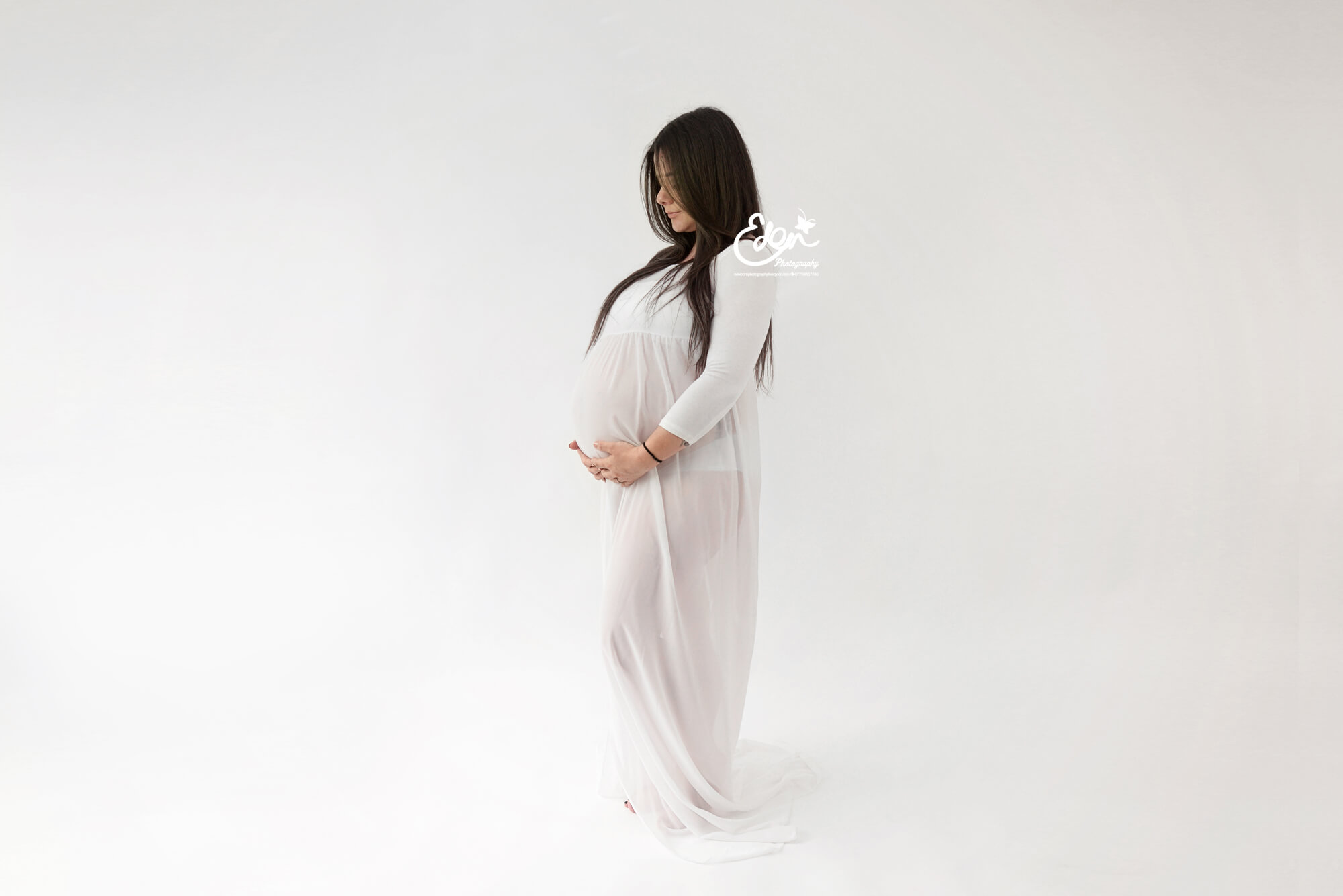 Pregnant lady looking down at her belly in her hands wearing a white flowing dress